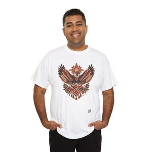May include: A white short-sleeved t-shirt featuring a central graphic of a stylized eagle with wings spread wide. The design incorporates geometric patterns, mountain motifs, and warm earth tones including shades of brown, orange, and cream.