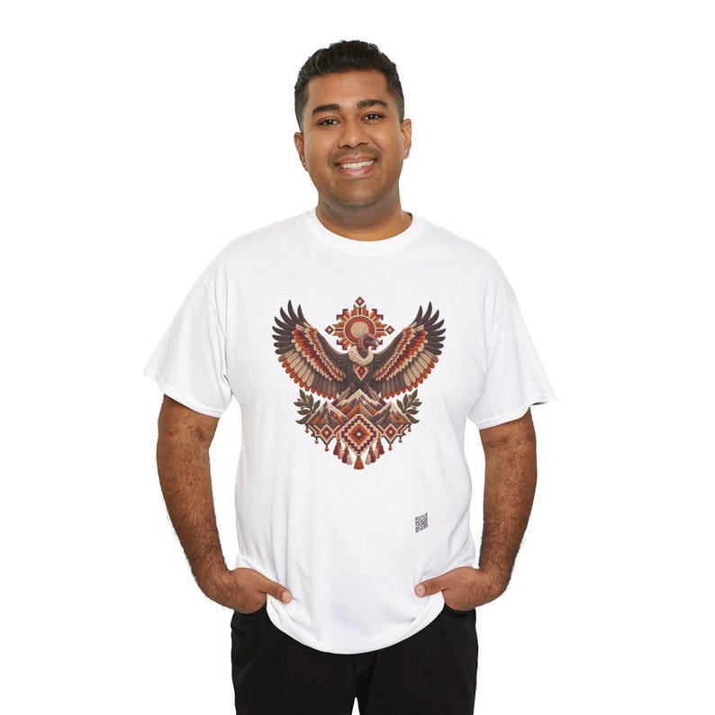 May include: A white short-sleeved t-shirt featuring a central graphic of a stylized eagle with wings spread wide. The design incorporates geometric patterns, mountain motifs, and warm earth tones including shades of brown, orange, and cream.