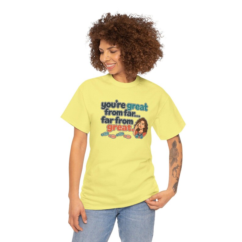 May include: A light yellow short-sleeve t-shirt featuring the text 'you're great from far... far from great.' in blue and red, accompanied by a small illustration of a person with curly hair and four colorful shoe icons.