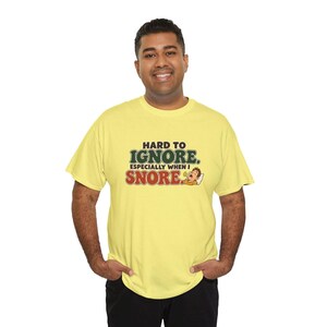 May include: A light yellow short-sleeved t-shirt featuring the text "HARD TO IGNORE. ESPECIALLY WHEN I SNORE." in dark green and red lettering, accompanied by a small cartoon illustration of a person sleeping and snoring.