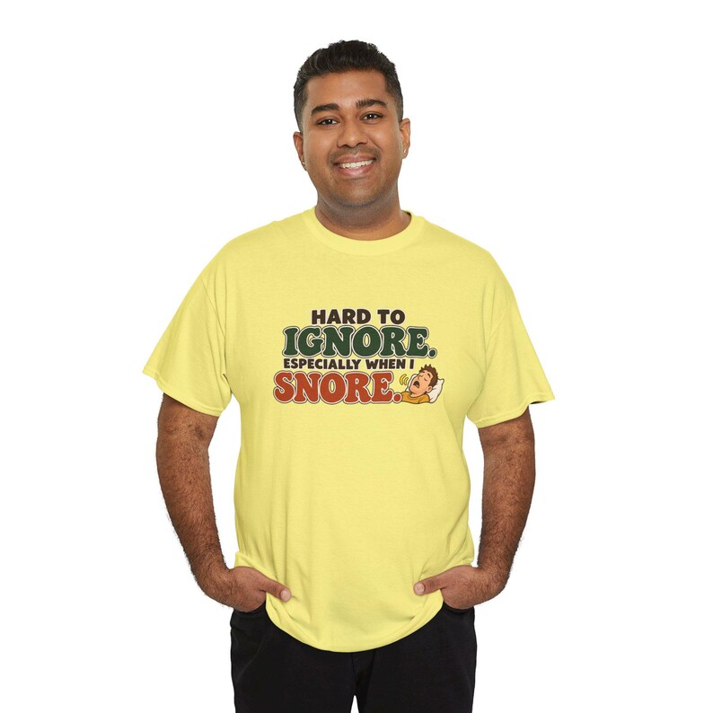 May include: A light yellow short-sleeved t-shirt featuring the text "HARD TO IGNORE. ESPECIALLY WHEN I SNORE." in dark green and red lettering, accompanied by a small cartoon illustration of a person sleeping and snoring.