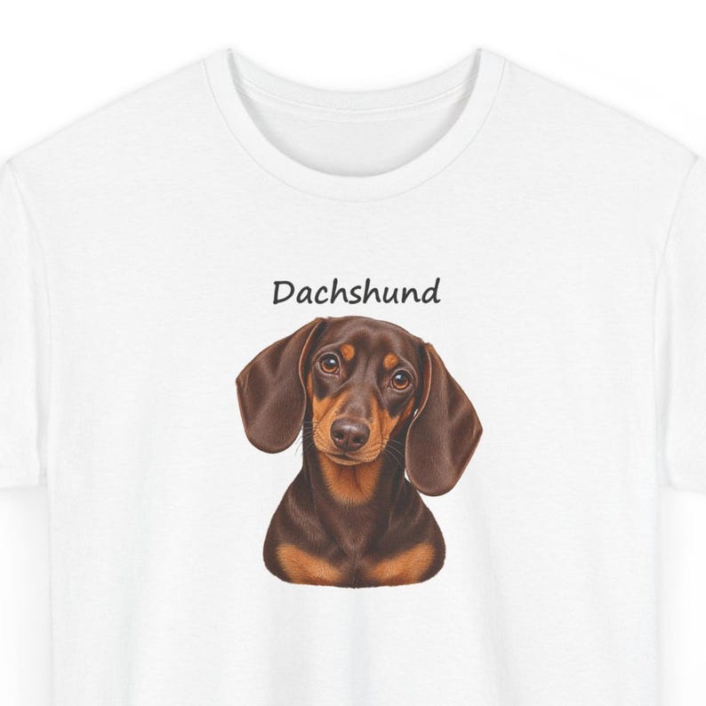 K&ouml;nnte beinhalten: Ein wei&szlig;es Kurzarm-T-Shirt mit einer zentrierten grafischen Illustration eines braun-schwarzen Dackels. &Uuml;ber dem Kopf des Hundes ist das Wort Dachshund in einer einfachen schwarzen serifenlosen Schrift gedruckt. Das Shirt hat einen Rundhalsausschnitt.