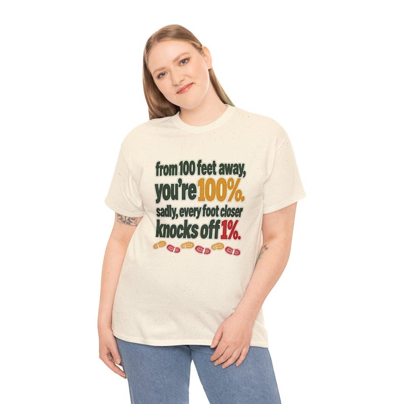 May include: A cream-colored short-sleeve t-shirt with dark green text that reads: from 100 feet away, you're 100%. sadly, every foot closer knocks off 1%. The text includes yellow and red accents with small footprint icons at the bottom.