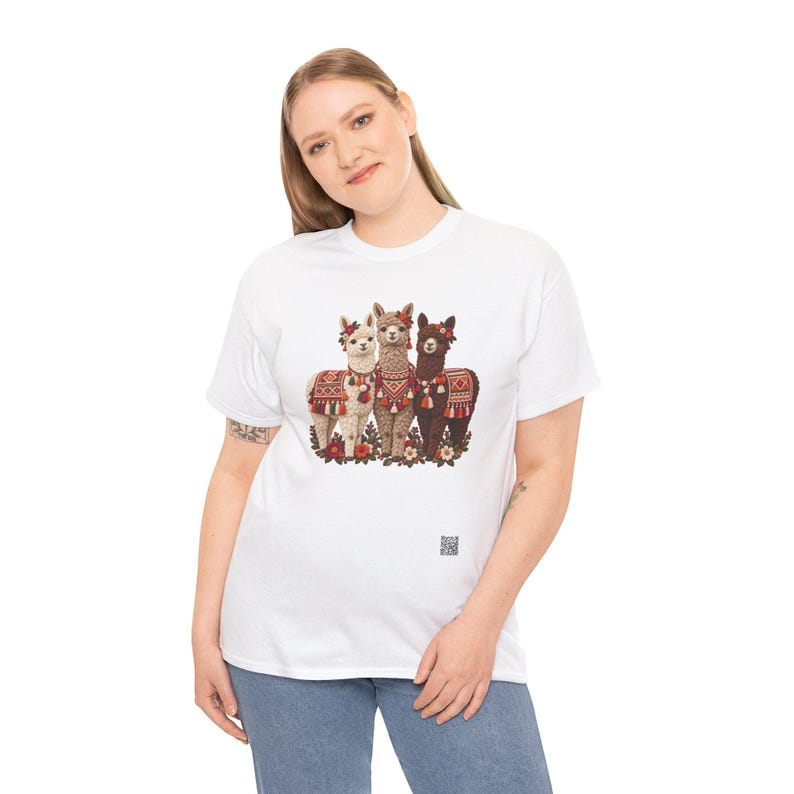 May include: A white short-sleeved t-shirt featuring a graphic print of three alpacas in the center. The alpacas are white, tan, and dark brown, each wearing colorful patterned blankets and floral headpieces. A small QR code is printed below the graphic.