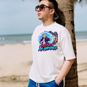 Może przedstawiać: Biały t-shirt z kr&oacute;tkim rękawem z nadrukiem graficznym przedstawiającym osobę w niebieskiej bańce energii oraz napisem "NOT IN MY AIRSPACE" pogrubionymi, kolorowymi literami. Wz&oacute;r zawiera fioletowy dym i stylizowany obiekt po prawej stronie.