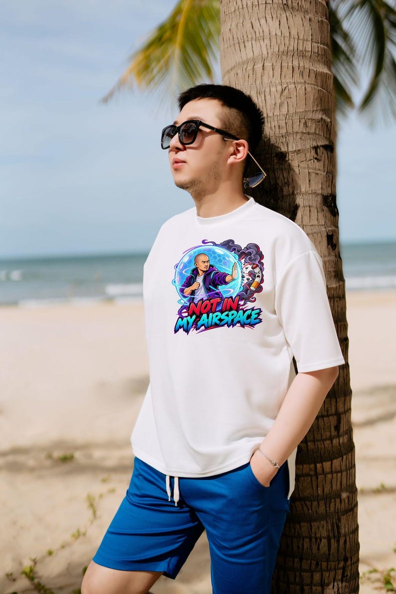 Może przedstawiać: Biały t-shirt z kr&oacute;tkim rękawem z nadrukiem graficznym przedstawiającym osobę w niebieskiej bańce energii oraz napisem "NOT IN MY AIRSPACE" pogrubionymi, kolorowymi literami. Wz&oacute;r zawiera fioletowy dym i stylizowany obiekt po prawej stronie.