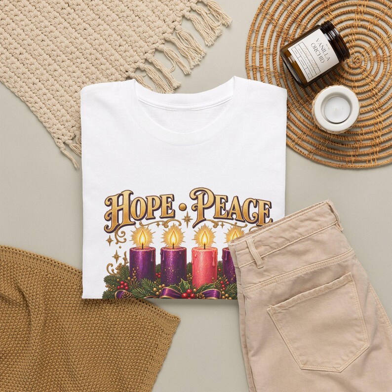 May include: A white t-shirt featuring a graphic print of four lit candles in purple and pink shades, surrounded by evergreen boughs and berries. Above the candles, the text reads HOPE &bull; PEACE in a decorative gold font with star accents.