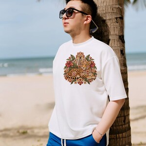 May include: A white short-sleeved t-shirt featuring a central graphic print of a leopard resting in front of a stepped pyramid, surrounded by tropical flowers and geometric patterns, paired with bright blue drawstring shorts.