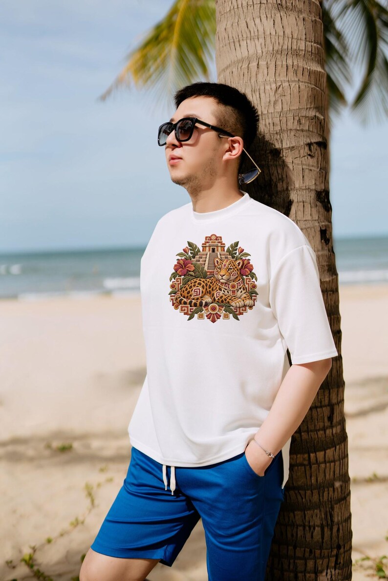 May include: A white short-sleeved t-shirt featuring a central graphic print of a leopard resting in front of a stepped pyramid, surrounded by tropical flowers and geometric patterns, paired with bright blue drawstring shorts.