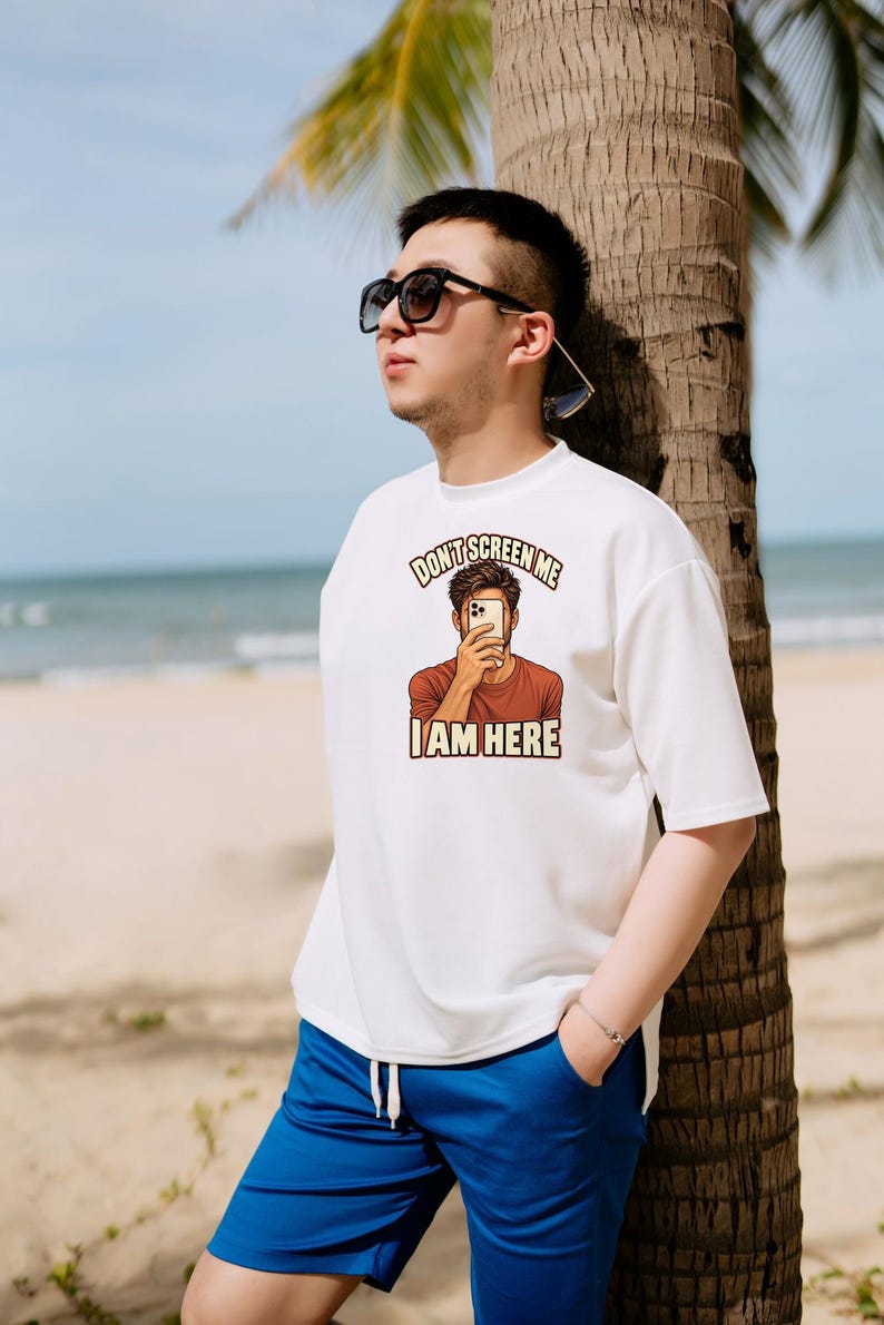 Op de afbeelding: Een wit T-shirt met korte mouwen en een grafische print van een persoon die een smartphone voor het gezicht houdt. De tekst boven de afbeelding luidt "DON'T SCREEN ME" en de tekst eronder "I AM HERE" in een vet, schreefloos lettertype.