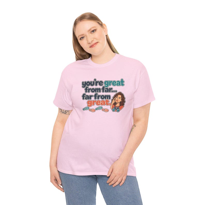 May include: A light pink short-sleeve t-shirt featuring a graphic print with the text "you're great from far... far from great." The design includes a cartoon illustration of a person looking through binoculars and small footprints.