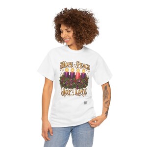 May include: A white short-sleeve t-shirt featuring a graphic of an Advent wreath with four lit candles, three purple and one pink, surrounded by greenery and ribbons. The text reads HOPE, PEACE, JOY, and LOVE in a decorative font.