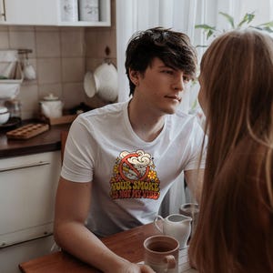 May include: A white short-sleeve t-shirt featuring a graphic print of a cigarette with a red prohibition symbol over it, surrounded by yellow and orange flowers, with the text "YOUR SMOKE IS NOT MY VIBE" in a bold, retro-style font.