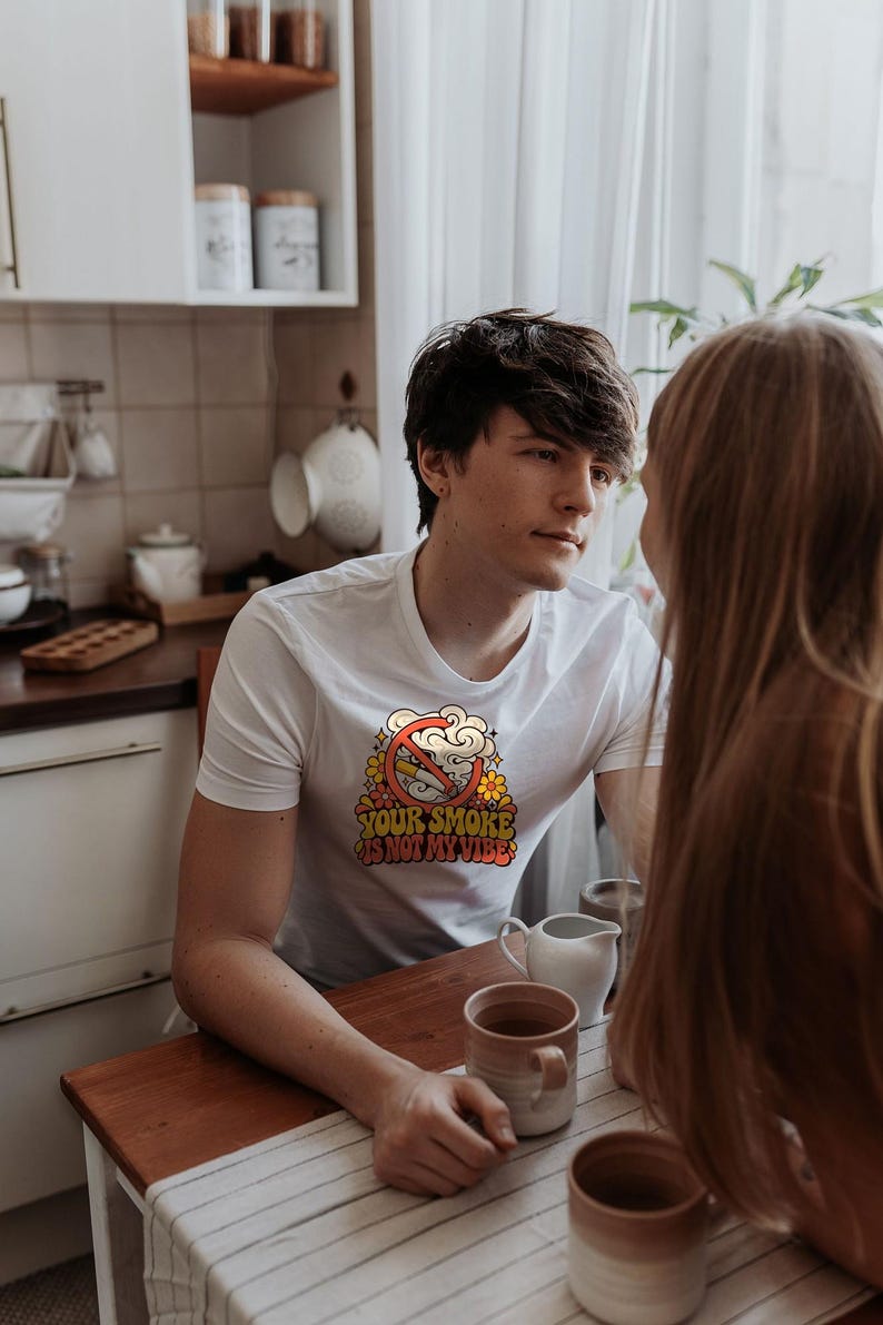 May include: A white short-sleeve t-shirt featuring a graphic print of a cigarette with a red prohibition symbol over it, surrounded by yellow and orange flowers, with the text "YOUR SMOKE IS NOT MY VIBE" in a bold, retro-style font.