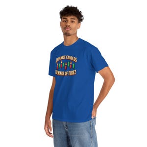 May include: A royal blue short-sleeve t-shirt featuring a graphic of red and green candlestick chart bars with flames. The text above the graphic reads "JAPANESE CANDLES" and the text below reads "BEWARE OF FIRE!" in a bold, arched font.