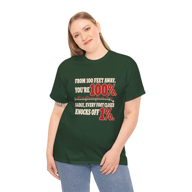 May include: A dark green short-sleeve t-shirt featuring the text "FROM 100 FEET AWAY, YOU'RE 100%. SADLY, EVERY FOOT CLOSER KNOCKS OFF 1%." The graphic includes a horizontal line with small red circular icons and a downward-pointing arrow.