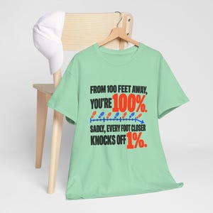 May include: A light green short-sleeve t-shirt featuring black and red text that reads: FROM 100 FEET AWAY, YOU'RE 100%. SADLY, EVERY FOOT CLOSER KNOCKS OFF 1%. The design includes a blue line graph with small red and blue footprint icons.
