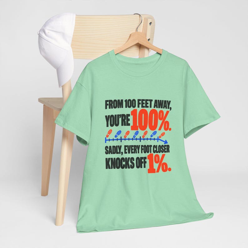 May include: A light green short-sleeve t-shirt featuring black and red text that reads: FROM 100 FEET AWAY, YOU'RE 100%. SADLY, EVERY FOOT CLOSER KNOCKS OFF 1%. The design includes a blue line graph with small red and blue footprint icons.