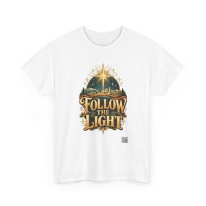May include: A white short-sleeve t-shirt featuring a central graphic of a glowing star above a landscape with the text FOLLOW THE LIGHT in gold lettering. The design includes a dark blue night sky, rolling hills, and a small QR code below.