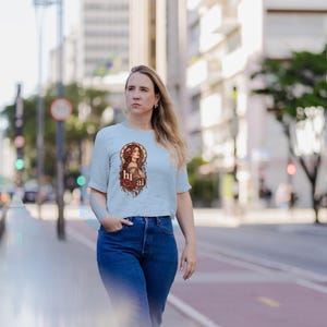 K&ouml;nnte beinhalten: Ein hellblaues kurz&auml;rmeliges T-Shirt mit einem Grafikdruck einer Frau mit langem braunem Haar, umgeben von roten Rosen und braunen Ornamenten, mit dem kleingeschriebenen wei&szlig;en Text "hi ai" in der Mitte des Designs.