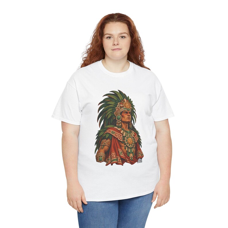May include: A white short-sleeved t-shirt featuring a graphic print of an Aztec warrior wearing a large green feathered headdress, a gold sun medallion necklace, and ornate red and gold patterned clothing with green accents.
