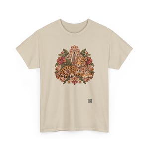 May include: A beige short-sleeve t-shirt featuring a central graphic of a spotted jaguar resting in front of a stepped pyramid. The design includes red hibiscus flowers, green leaves, and geometric patterns in shades of orange and brown.