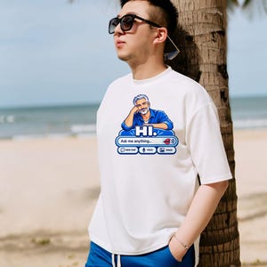 May include: A white short-sleeved t-shirt featuring a graphic of a man in a blue shirt with the text HI. Ask me anything. The graphic includes icons for new chat, voice, and image, along with a red lip icon and a sound wave symbol.