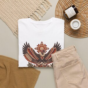 May include: A white t-shirt featuring a graphic print of a condor with wings spread wide, positioned below a geometric sun symbol and above mountain peaks, rendered in shades of brown, orange, and cream with intricate patterns.