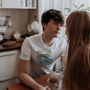 May include: A white short-sleeved t-shirt featuring a graphic of a fish jumping through blue water waves with the text "CURRENTLY SWIMMING AGAINST THE CURRENT" printed in a bold, stylized font across the chest area of the garment.