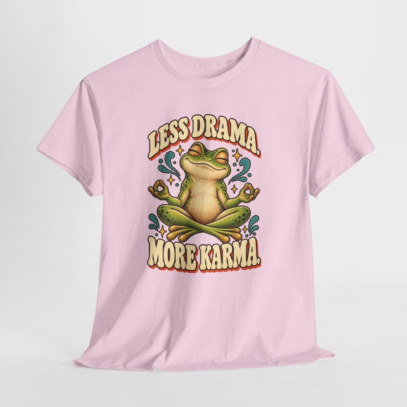 May include: A light pink short-sleeve t-shirt featuring a graphic of a green frog sitting in a lotus position with eyes closed. The text above the frog reads LESS DRAMA and the text below reads MORE KARMA in a bold, retro-style font.