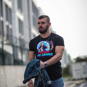 Może przedstawiać: Czarny t-shirt z kr&oacute;tkim rękawem z nadrukiem graficznym przedstawiającym osobę w niebieskiej bańce energetycznej blokującą papierosa. Napis "NOT IN MY AIRSPACE" jest wydrukowany pogrubionymi czerwonymi i niebieskimi literami pod grafiką.