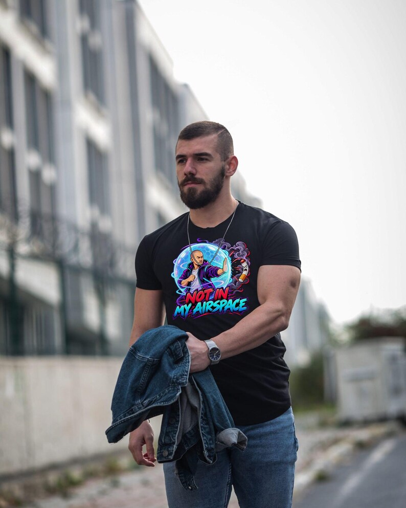Może przedstawiać: Czarny t-shirt z kr&oacute;tkim rękawem z nadrukiem graficznym przedstawiającym osobę w niebieskiej bańce energetycznej blokującą papierosa. Napis "NOT IN MY AIRSPACE" jest wydrukowany pogrubionymi czerwonymi i niebieskimi literami pod grafiką.