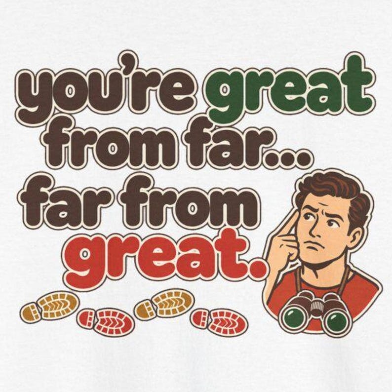 May include: A white graphic t-shirt featuring the text you're great from far... far from great. in brown, green, and red. An illustration of a person with brown hair, a red shirt, and binoculars around their neck is included.
