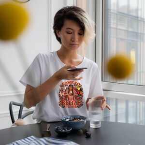 K&ouml;nnte beinhalten: Ein wei&szlig;es kurz&auml;rmeliges T-Shirt mit einem farbenfrohen Grafikdruck auf der Brust. Das Design zeigt eine Person in traditioneller Kleidung mit dem Text "COME TO BELIZE PLEASE", umgeben von Blumen- und Tropenvogelmotiven.