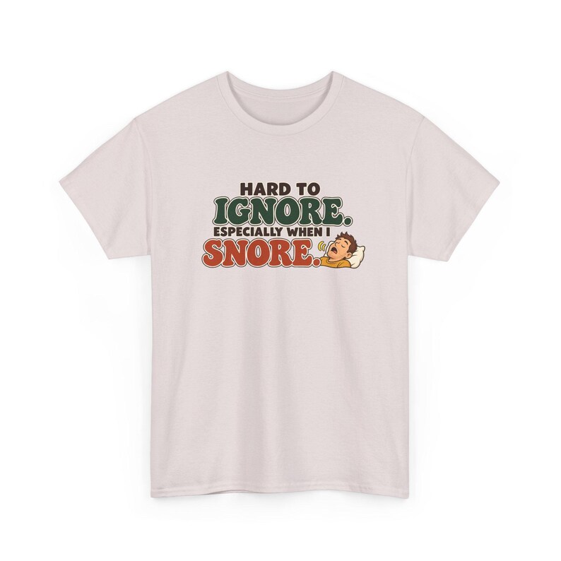 May include: A light pink short-sleeve t-shirt featuring the text "HARD TO IGNORE. ESPECIALLY WHEN I SNORE." in dark green and red lettering, accompanied by a cartoon illustration of a person sleeping on a pillow with a speech bubble.
