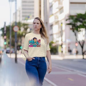May include: A cream-colored short-sleeve t-shirt featuring a graphic print of a person with dark hair resting their chin on their hand. The text reads "hi AI" and "ASK ME ANYTHING" with small icons below in pink, blue, and orange boxes.
