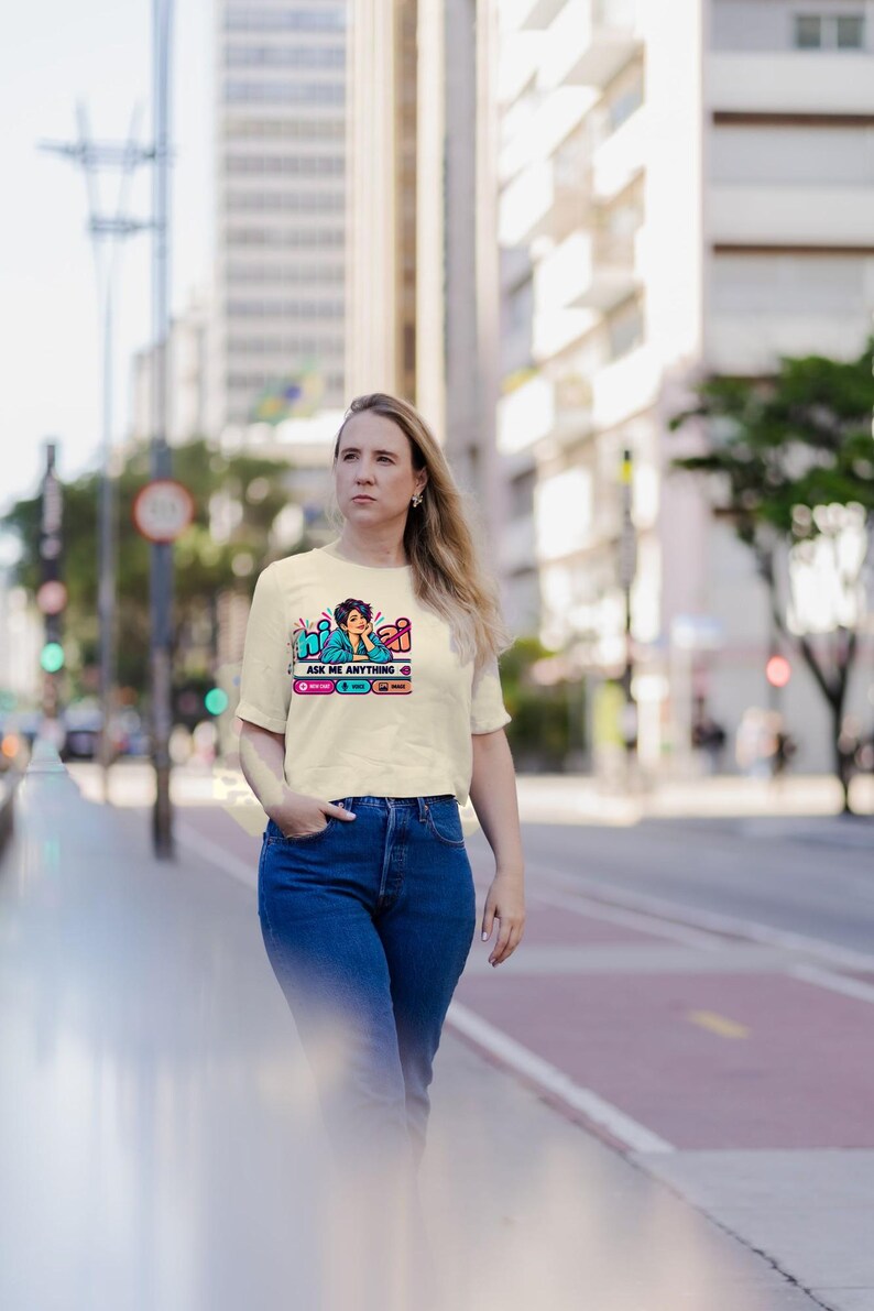 May include: A cream-colored short-sleeve t-shirt featuring a graphic print of a person with dark hair resting their chin on their hand. The text reads "hi AI" and "ASK ME ANYTHING" with small icons below in pink, blue, and orange boxes.