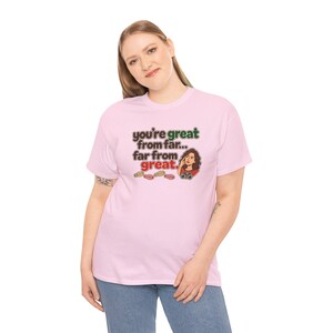 May include: A light pink short-sleeve t-shirt featuring the text you're great from far... far from great. The design includes a cartoon illustration of a person looking through binoculars and small colorful footprint icons below the text.