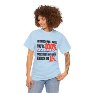 May include: A light blue t-shirt featuring black and red text that reads: FROM 100 FEET AWAY, YOU'RE 100%. SADLY, EVERY FOOT CLOSER KNOCKS OFF 1%. The design includes a horizontal line with small red and blue footprint icons.