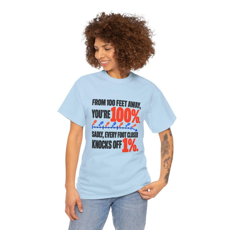 May include: A light blue t-shirt featuring black and red text that reads: FROM 100 FEET AWAY, YOU'RE 100%. SADLY, EVERY FOOT CLOSER KNOCKS OFF 1%. The design includes a horizontal line with small red and blue footprint icons.