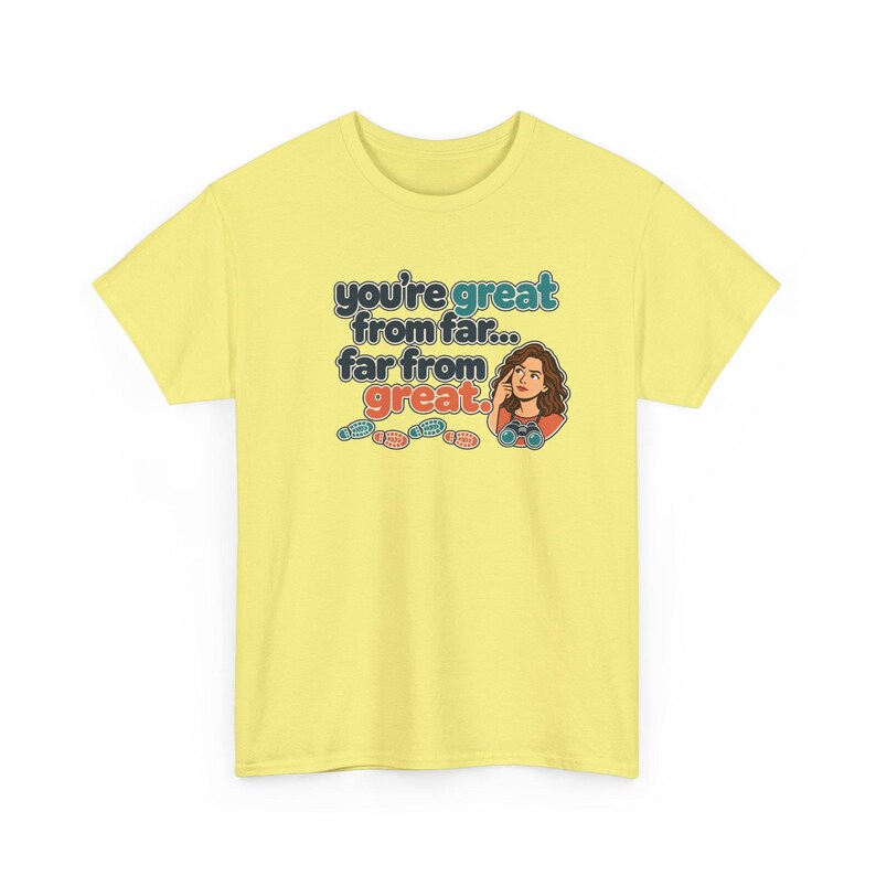 May include: A pale yellow short-sleeve t-shirt featuring a graphic of a person looking through binoculars with the text "you're great from far... far from great." and four small colorful footprints printed below the text.