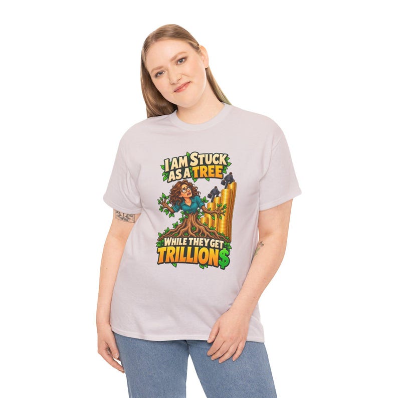 May include: A light gray t-shirt featuring a graphic of a person with brown hair and glasses whose lower body is transformed into a tree trunk. The text reads I AM STUCK AS A TREE WHILE THEY GET TRILLIONS, with gold coins stacked nearby.
