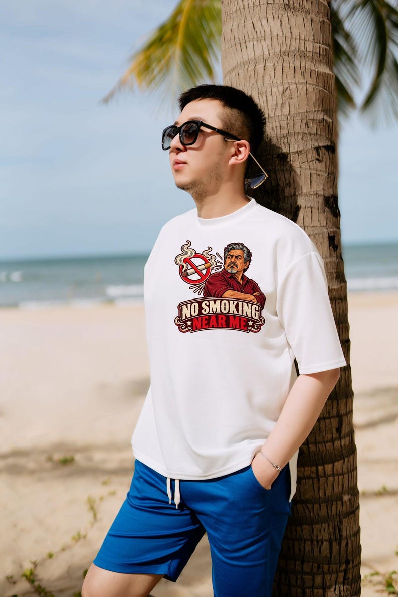 May include: A white short-sleeved t-shirt featuring a graphic of a man with a mustache and a red no-smoking symbol above him. The text "NO SMOKING NEAR ME" is printed in bold red and black lettering below the illustration on the chest.