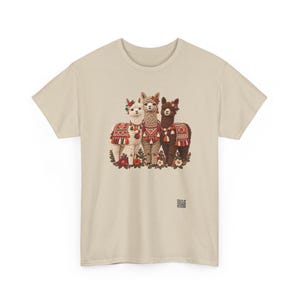 May include: A beige short-sleeve t-shirt featuring a graphic of three llamas in white, tan, and dark brown. The llamas are adorned with colorful patterned blankets, tassels, and floral headpieces, standing amidst a cluster of flowers.