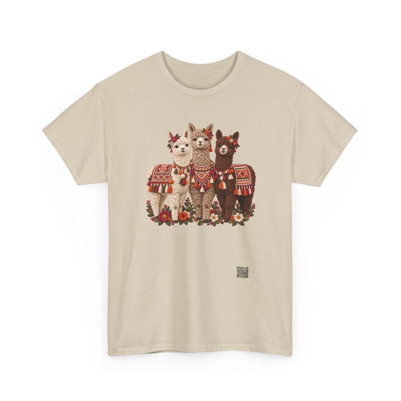 May include: A beige short-sleeve t-shirt featuring a graphic of three llamas in white, tan, and dark brown. The llamas are adorned with colorful patterned blankets, tassels, and floral headpieces, standing amidst a cluster of flowers.