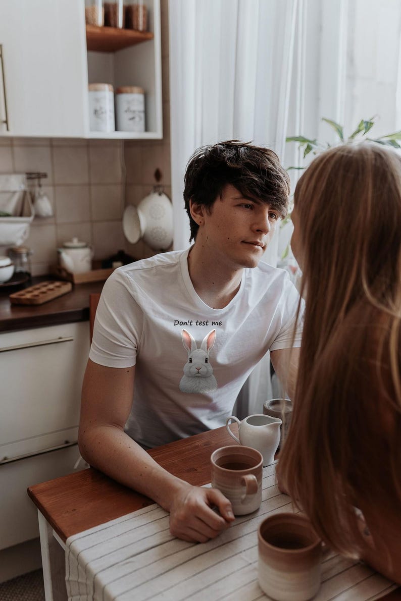 May include: A white short-sleeved t-shirt featuring a graphic of a gray rabbit centered on the chest. Above the rabbit, the text "Don't test me" is printed in a simple black font. The shirt has a crew neckline and a standard fit.