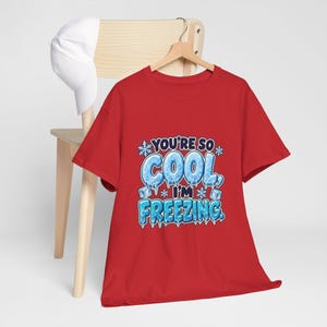 May include: A red short-sleeved t-shirt featuring a graphic with the text "YOU'RE SO COOL, I'M FREEZING." The lettering is designed to look like ice, accompanied by illustrations of snowflakes and ice cubes in shades of blue and white.