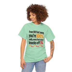 May include: A light green short-sleeve t-shirt featuring the black text: from 100 feet away, you're 100%. sadly, every foot closer knocks off 1%. The numbers 100% and 1% are in orange and red, with small colorful footprint icons below.
