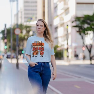 May include: A light blue short-sleeve t-shirt featuring the text I AM MEDIUM LOOKING FOR RARE in orange and red letters, accompanied by cartoon illustrations of a steak and a piece of meat with a magnifying glass.