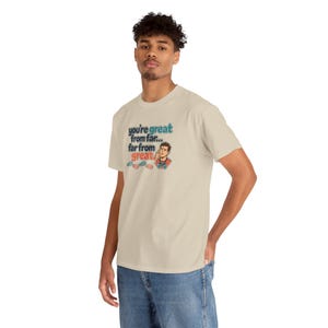 May include: A beige short-sleeve t-shirt featuring a graphic of a person looking through binoculars. The text reads, you're great from far... far from great. The design includes small blue and red binocular icons below the text.